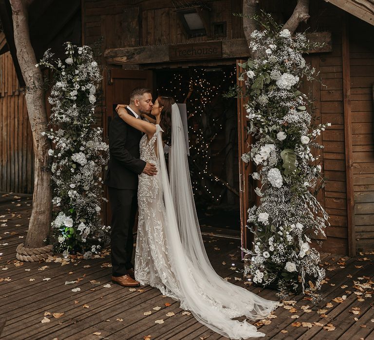 The bride and groom share a kiss at the entrance to Alnwick Treehouse with white flower column arrangements 