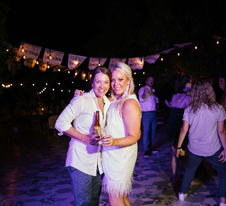 Blonde bride wears short reception dress finished with feathered edging as she stands beside her bride in white shirt and blue trousers during outdoor reception in Mexico