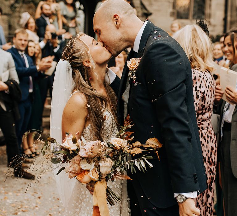 The bride in a leaf patterned wedding dress kissing the groom in a navy suit at the end of their confetti exit 