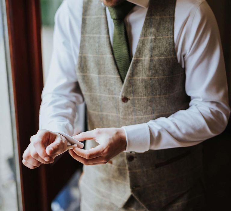 Groom in white shirt wearing a light green checkered trousers and waistcoat with dark green tie putting on cufflink accessories 