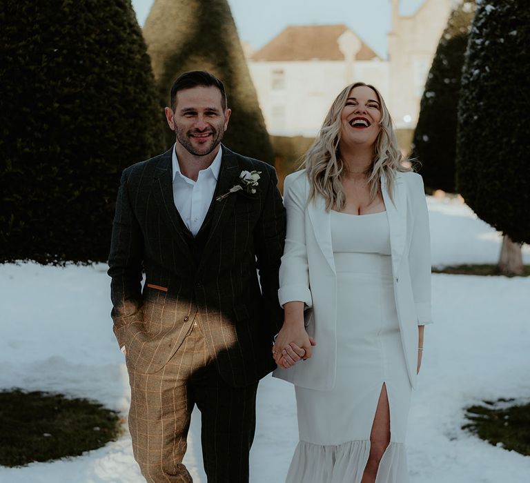 Winter Christmas wedding with the groom in dark grey checkered suit with the bride in split leg wedding dress, blazer and pointed shoes walking in the snow 