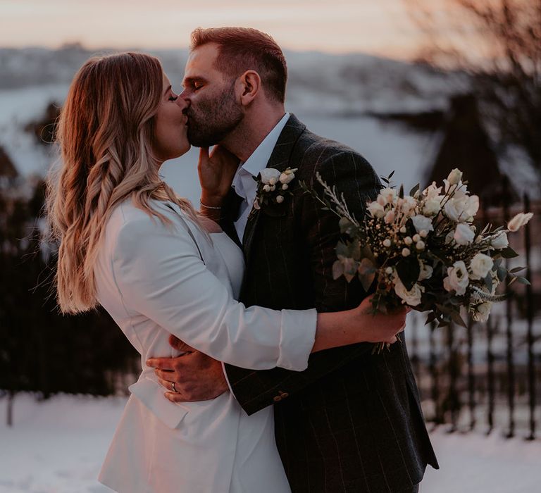 Bride with curled hair in a white blazer holding a white flower bouquet kisses the groom in a dark grey checkered suit during the winter sunset 