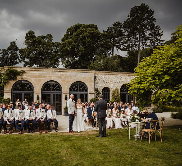 The bride and groom stand together with the celebrant and the rest of the wedding guests seated for outdoor ceremony 