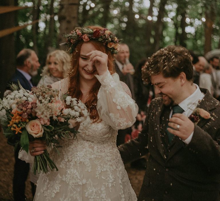 Bride wearing off the shoulder long sleeve boho wedding dress with flower crown walking with the groom as confetti is thrown over the guests 