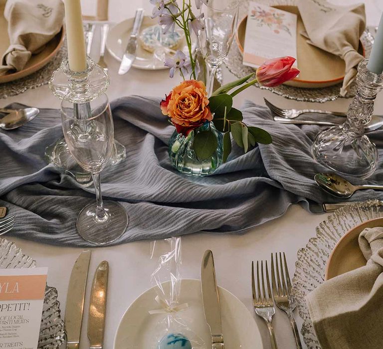 Cream tapered candle in glass candlestick holder on cornflower blue wedding table runner with bright blue glass bottles with dried flowers