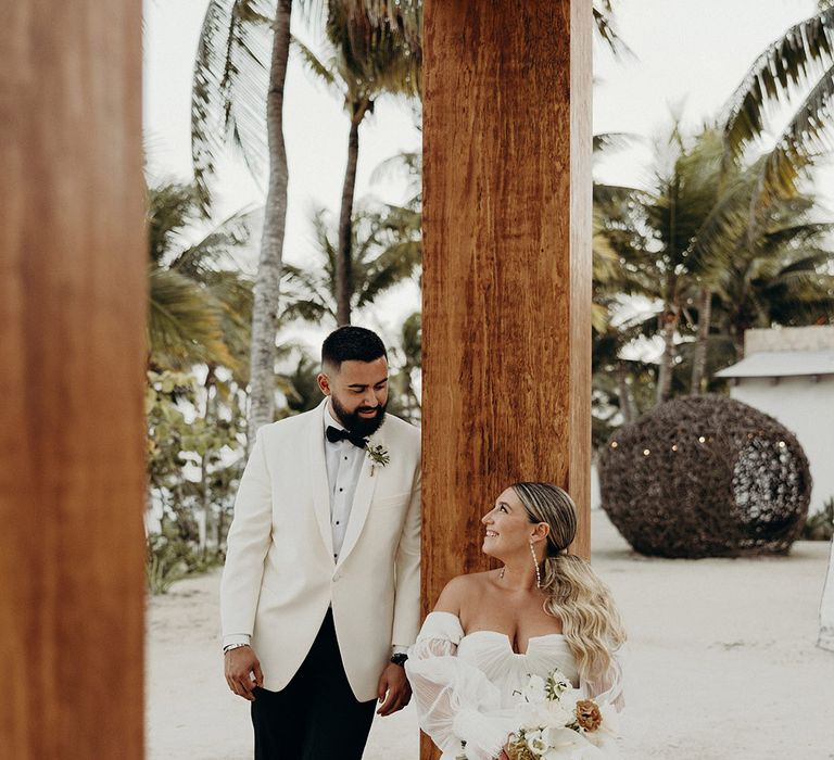 modern blue venado beach wedding with groom in a white tuxedo jacket and bride in a strapless tulle wedding dress with sheer long sleeves sitting on a wooden frame