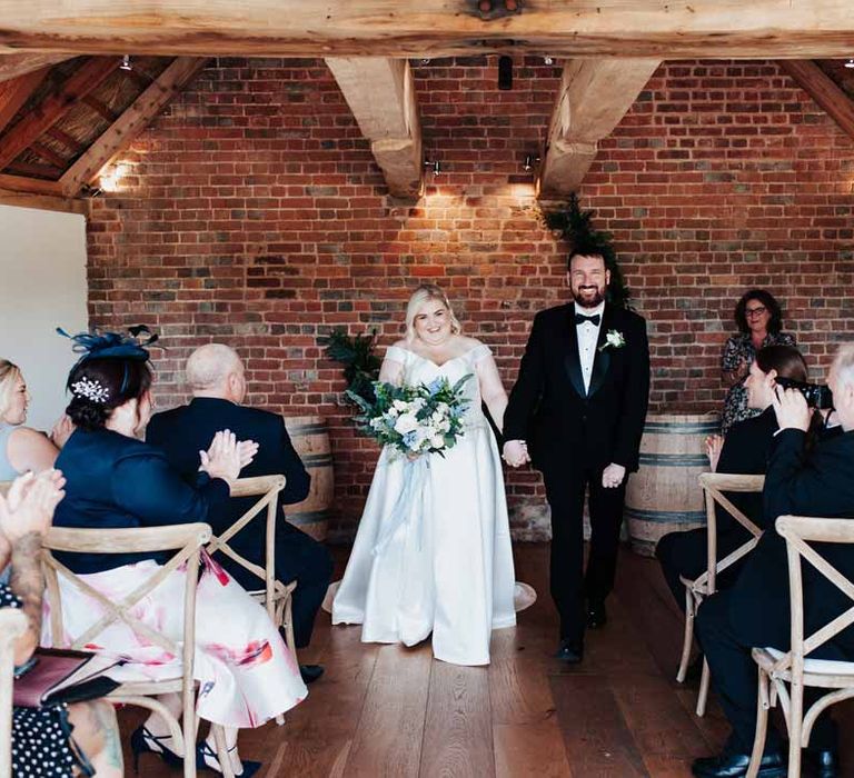 Bride in satin off the shoulder wedding dress holding eucalyptus, foliage, garden rose, gardenia, bluebell and dried flower bridal bouquet tied with dusky blue ribbon walking back down the aisle with groom in classic black grooms three piece tuxedo with white rose boutonniere and black bowtie 