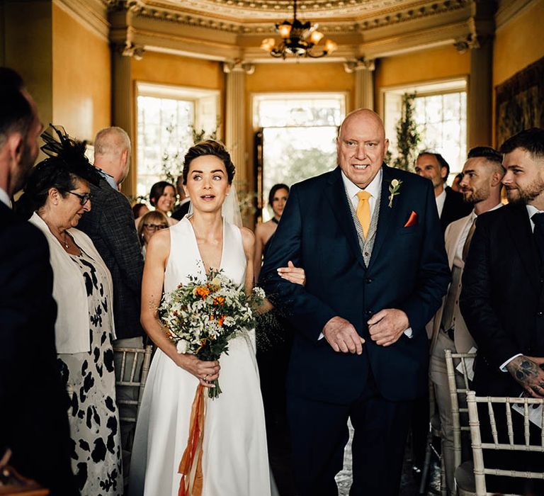 Father of the bride in a dark blue suit with yellow tie walks the bride down the aisle to meet the groom 