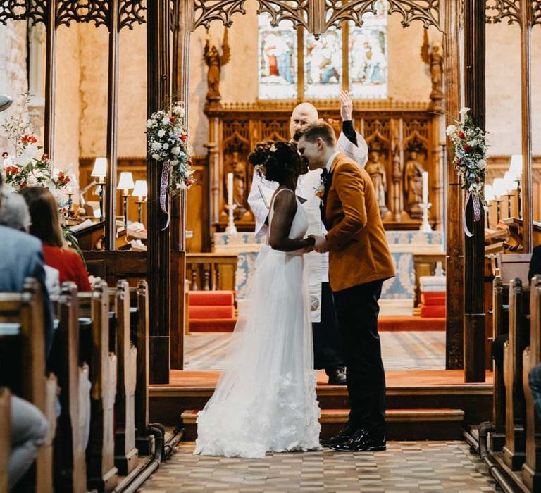 Bride in v-neck sleeveless wedding dress with 3D floral applique train kissing groom in crushed burnt orange velvet grooms blazer, black bowtie and black suit trousers at the alter of Brinsop Court wedding venue