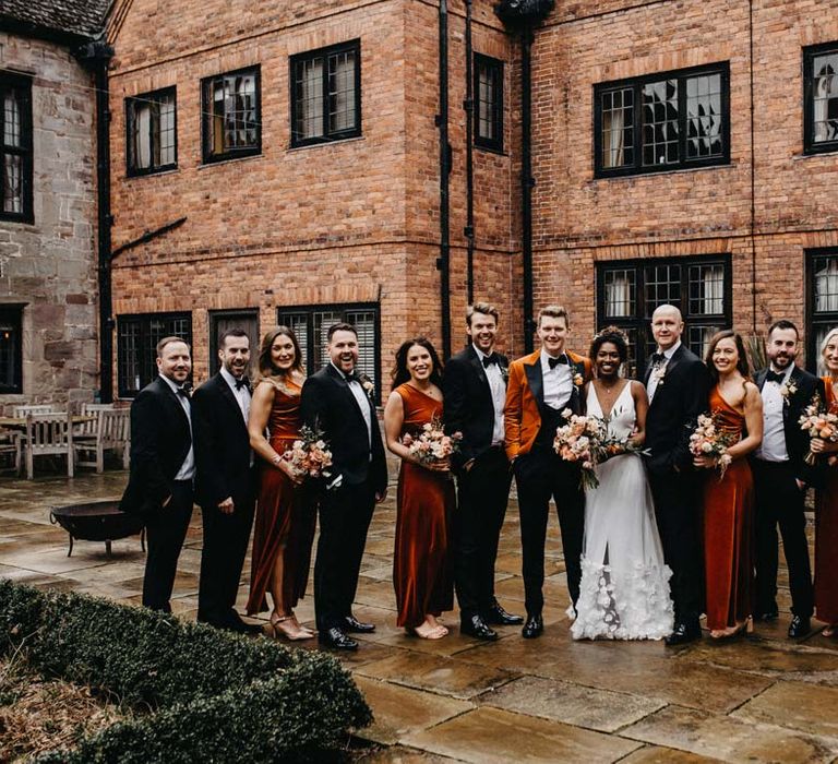 Wedding party wearing classic black groomsmen suits, one shoulder burnt orange bridesmaid dresses, crushed burnt orange velvet grooms blazer, black bowtie and black suit trousers and a v-neck sleeveless wedding dress with 3D floral applique train all standing outside of Brinsop Court wedding venue