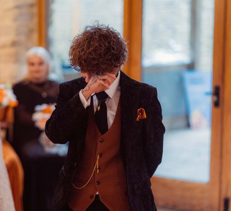 Bride in dark velvet bridal jacket with rast waistcoat and pocket square, black suit trousers `nd gold pocket watch wiping away a tear at the alter of Kingscote Barn Tetbury