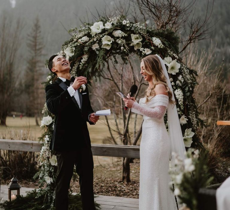 Bride in off shoulder sparkly mermaid wedding dress reading funny wedding speech to groom in classic black tuxedo, black bowtie and white garden rose boutonniere standing by white garden rose, eucalyptus, baby's-breath and foliage wedding arch at French destination ski wedding