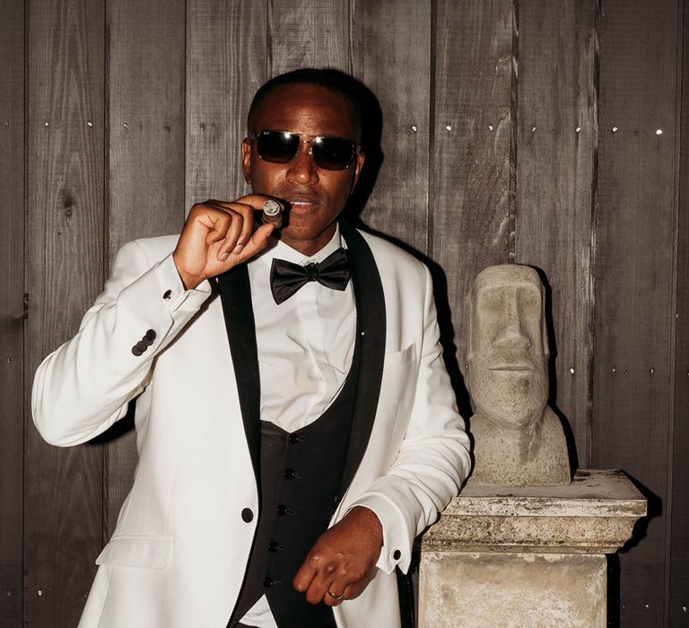 Groom in white and black tuxedo and sunglasses smoking a cigar at black-tie wedding