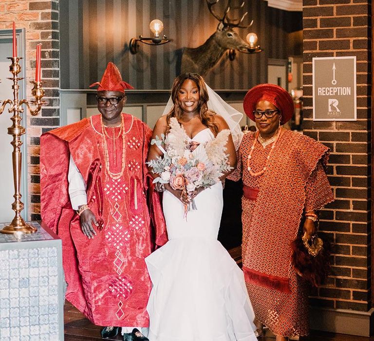 Bride in a fishtail mermaid wedding dress with her parents in red Yoruba wedding outfits 