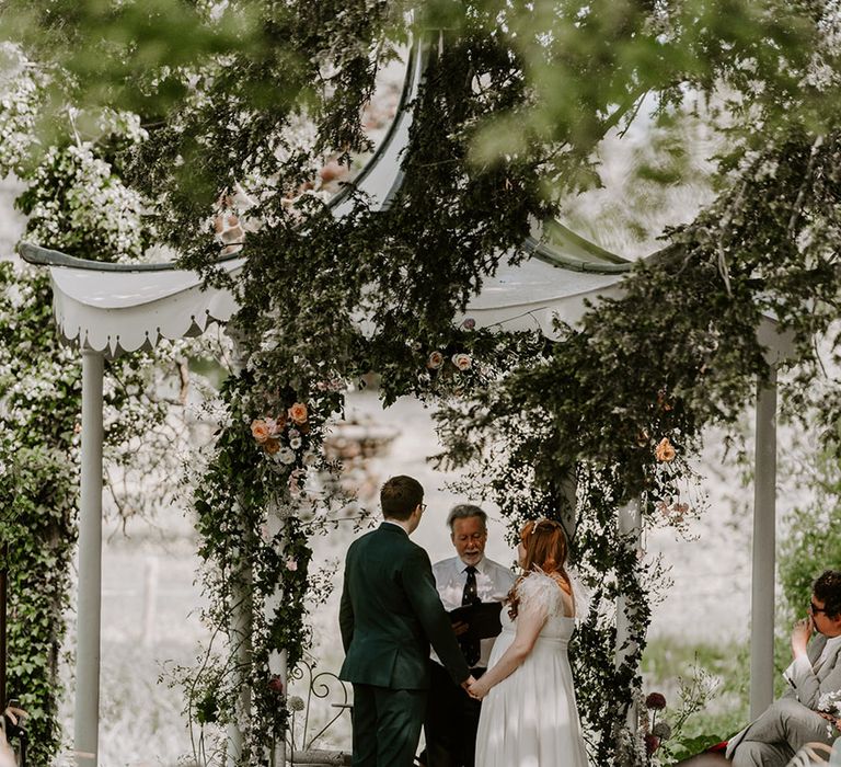 The bride and groom hold hands for their outdoor wedding ceremony with neutral wedding flowers decorating the altar space 