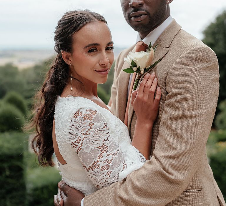 Groom in beige suit standing with the bride in a boho lace wedding dress with long sleeves 