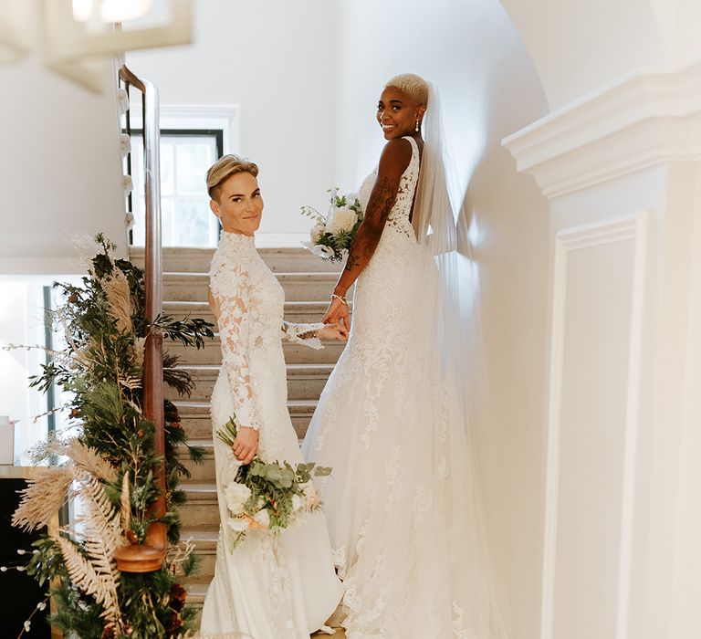 Two brides in lace wedding gowns holding hands with neutral wedding bouquets and neutral wedding decorations on the staircase 