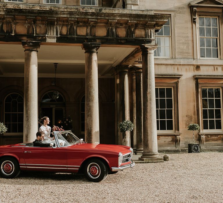 Vintage red wedding car being driven by groom with the bride sitting in the passenger seat 