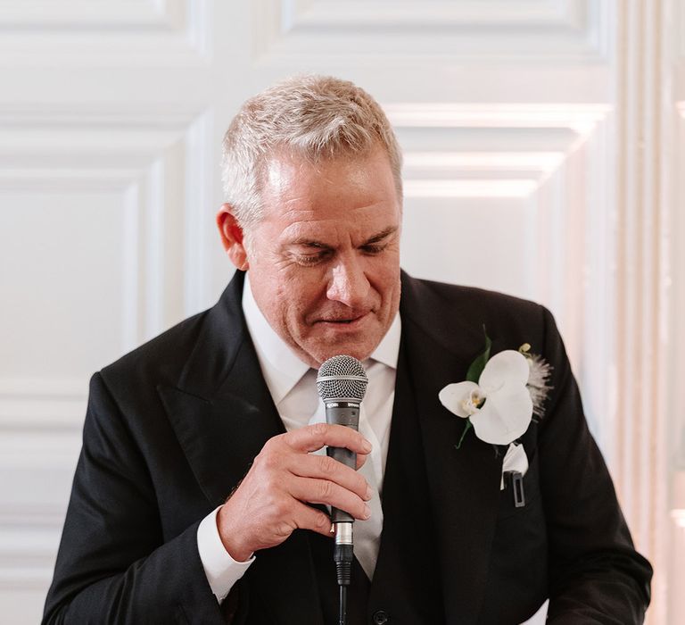 Groom in black wedding suit with white orchid buttonhole reading wedding speech 