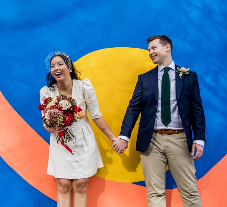 Portrait of a bride in a shirt wedding dress holding a red flower bouquet and her grooms hand standing in front of a colourful graffiti wall