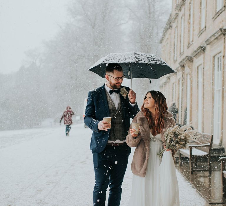 Bride in fur coat and groom in navy suit hold cups whilst walking under black umbrella during a snowfall at Cannon Hall