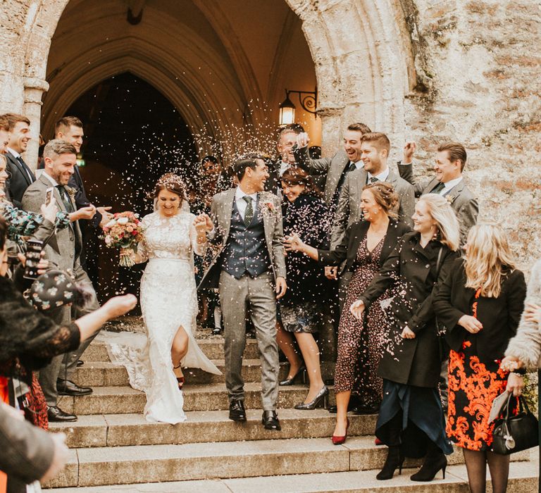 Church exit wedding confetti moment with the bride in a lace Hermione de Paula wedding dress with front split and the groom in a three-piece grey and navy wool suit