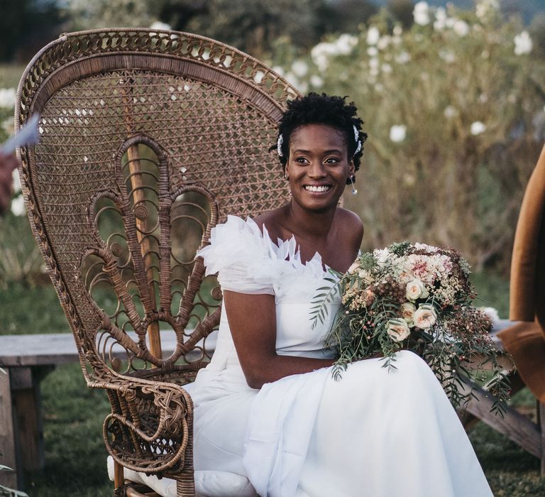 West African bride in a Georges Hobeika wedding dress with feather shoulder detail sitting on a wicker chair at her outdoor wedding ceremony 