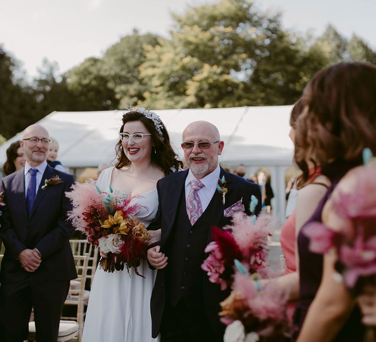 Bride in white cat eye glasses, bridal headband and Rime Arodaky wedding dress holds white and pink rose and pampas grass wedding bouquet walking accompanied up the aisle