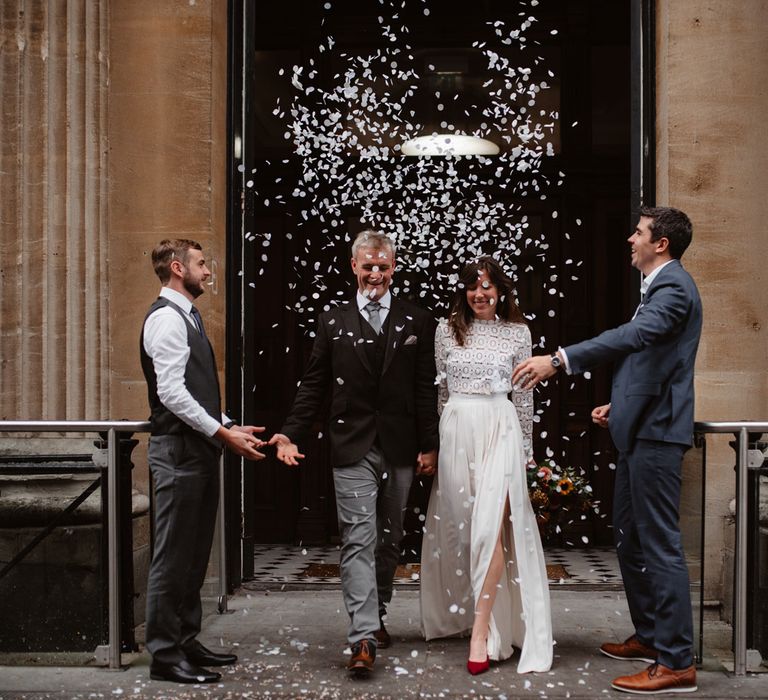 Bride in long sleeved lace top Self Portrait wedding dress holding colourful bouquet stands walks through confetti thrown by guests holding hands with groom in brown woollen blazer and waistcoat at Bristol Registry Office