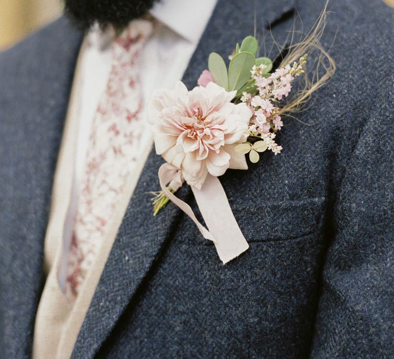 Groom wears a pink lapel flower with brown twine against a blue tweed blazer with a pink tie for English garden party wedding