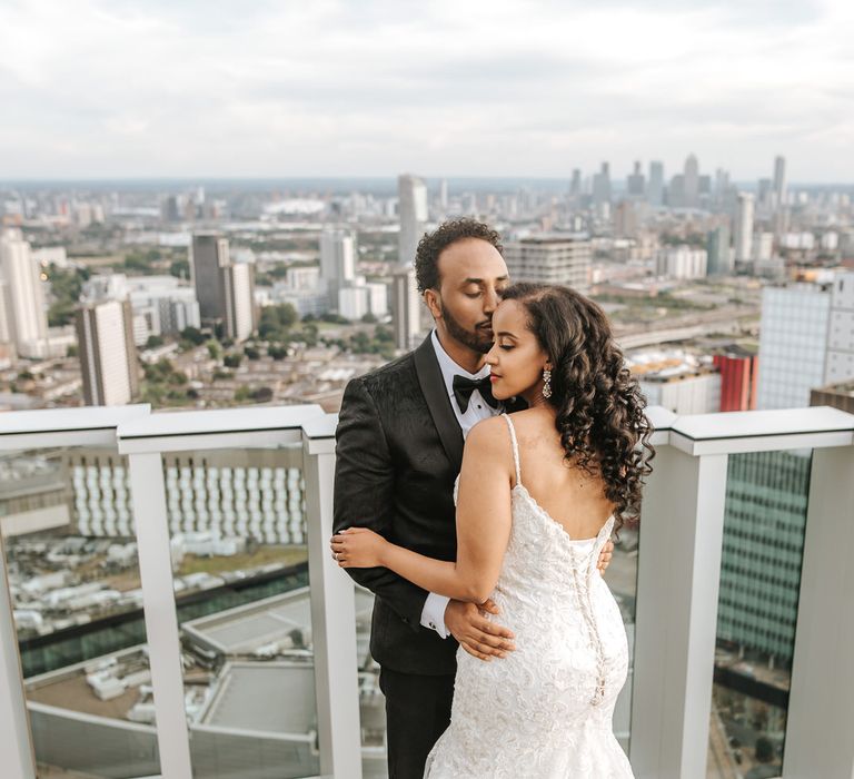 Groom in a black jacquard tuxedo jacket kissing his brides head in a princess fitted lace wedding dress with corset back 