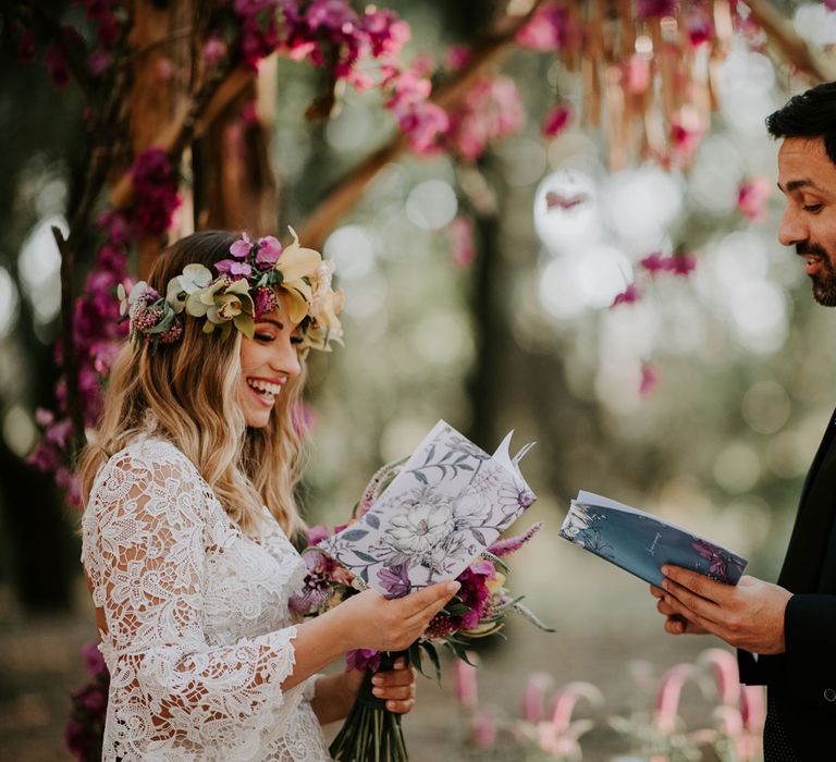 Bride in long sleeved lace boho wedding dress and flower crown reading does with groom in navy suit and bow tie in front of wooden wedding arch decorated with pink flowers