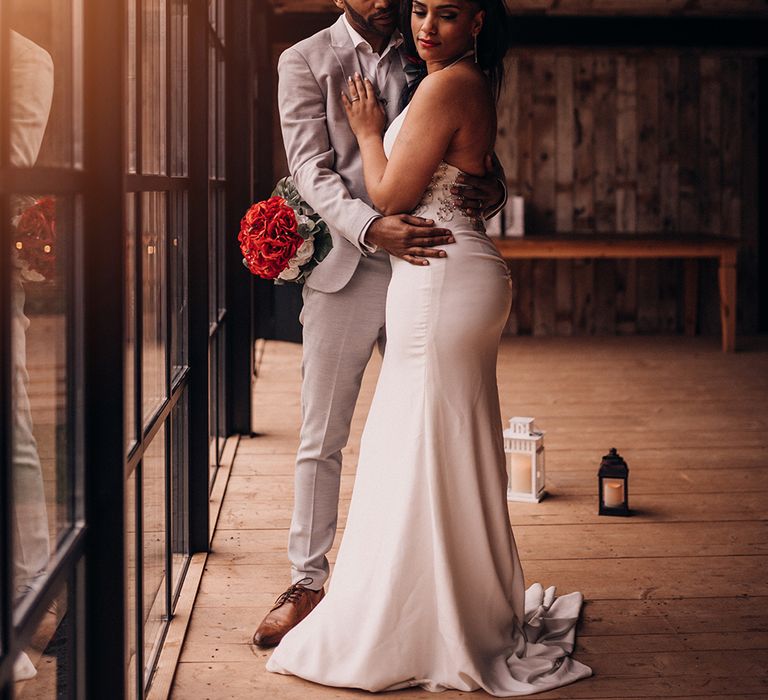 Groom wraps arms around bride as he looks at her, and bride holds bouquet of roses 