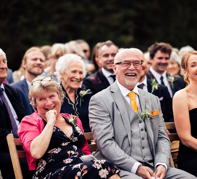 Wedding guests including father of the bride in grey suit and maid of honour in black strapless jumpsuit sit laughing at for lakeside wedding ceremony