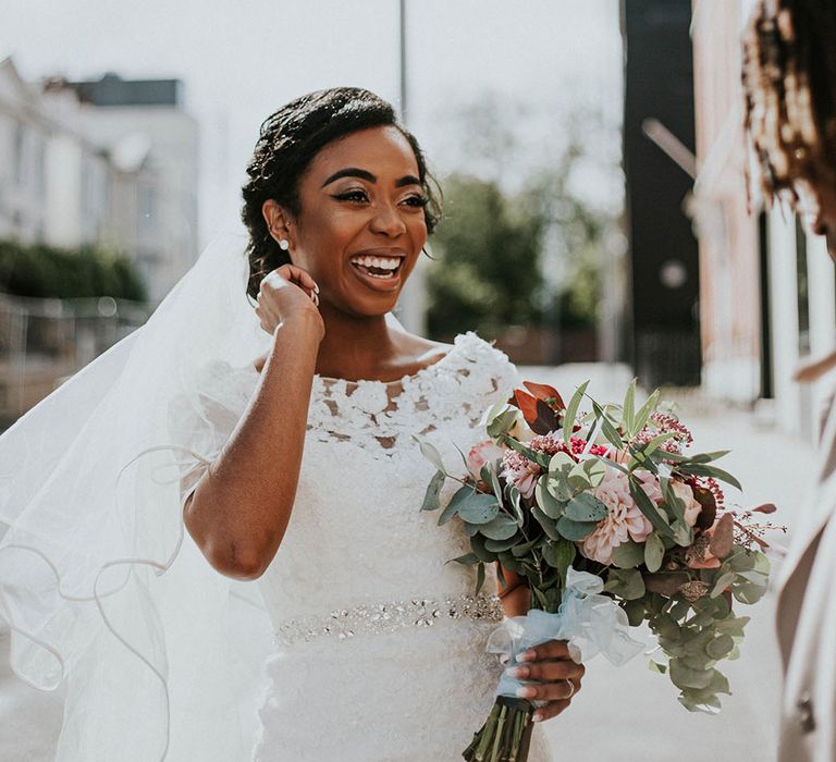 Beautiful bride in a fitted lace wedding dress with belt detail laughing and holding a pink and red wedding bouquet with eucalyptus