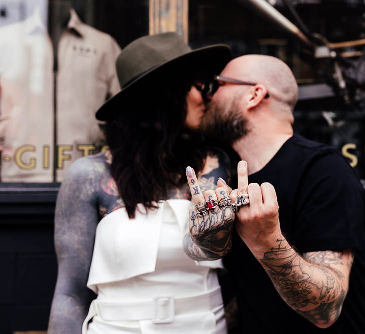 Tattooed bride and groom holding their wedding fingers up whilst kissing 