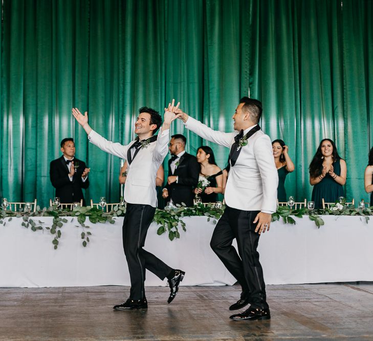 A gay couple wearing matching white tuxedo jackets enter their wedding breakfast. They walk past the top table to cheers and clapping. Photography by Beatrici Photography. 