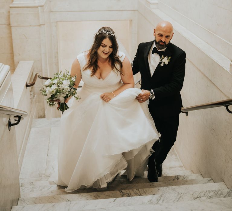 Bride walks up the steps on her wedding day as wedding party helps her lift skirt of wedding gown