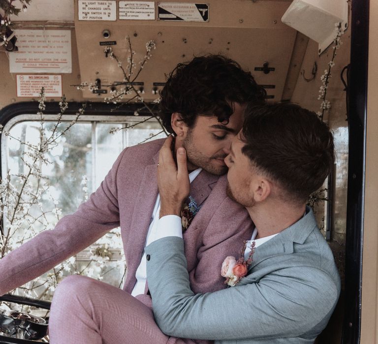 Groom sits on groom whilst wearing pastel suits and kissing on wedding day