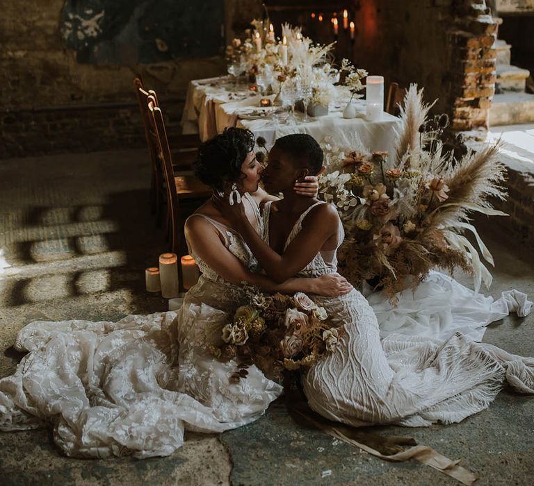 Two brides in an embellished wedding dress and appliqué gown kissing on the floor next to their wedding reception table at The Asylum wedding venue 