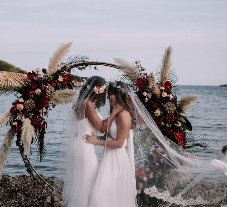 Two brides under one veil getting married on the beach in front of flower installation made of red anthuriums, roses, pampas grass, dried hydrangea, pink carnations, eucalyptus and palm leaves