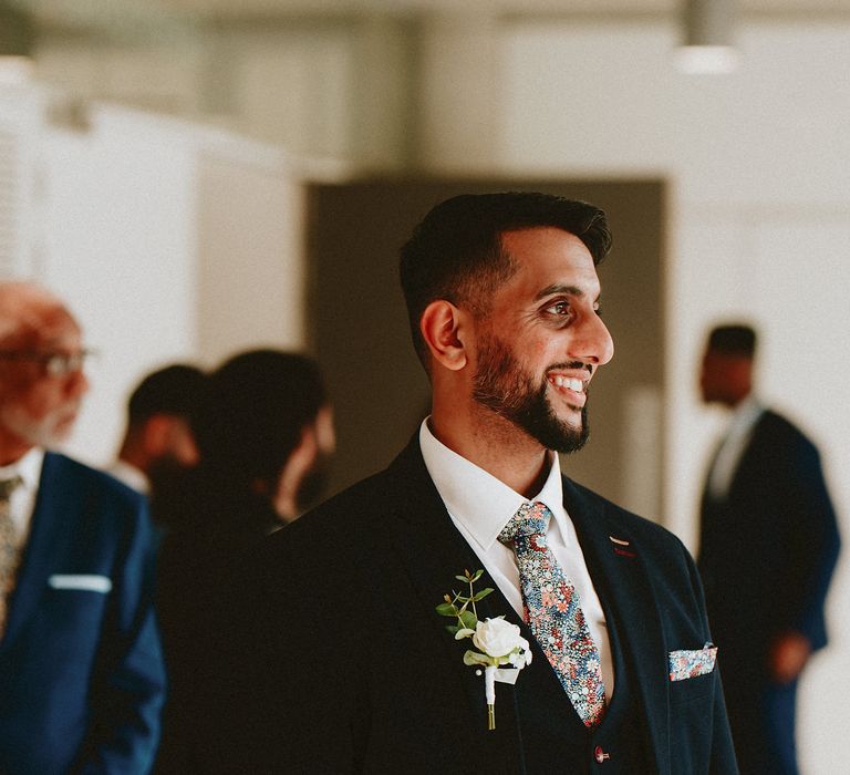 Stylish groom in a navy blue three-piece suit with floral tie smiling as his bride walks down the aisle 