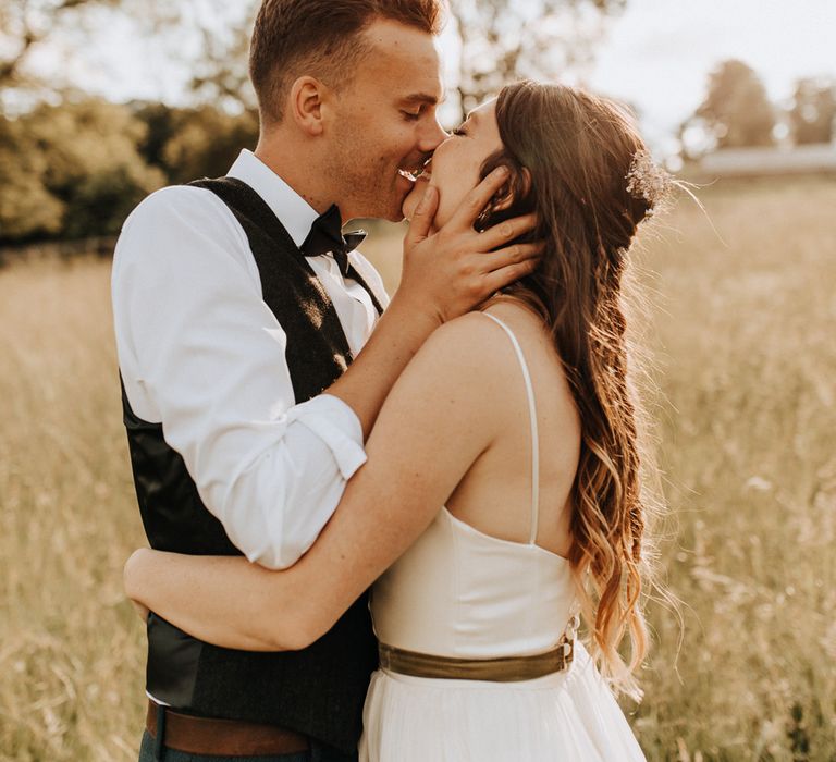 Bride in white cami wedding dress with gold ribbon waistband hugs groom in white shirt, waistcoat and bow tie as they kiss in field at garden wedding reception