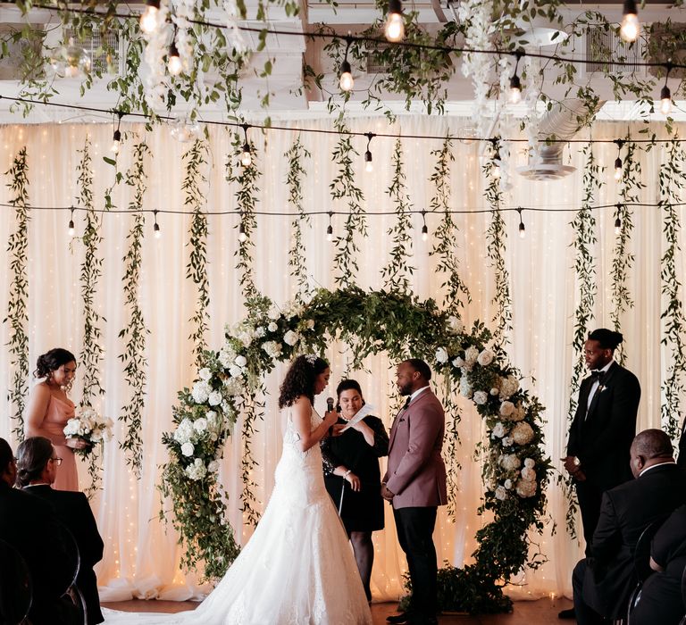 Bride & groom during wedding ceremony in front of floral archway and curtain whilst guests are seated on clear chairs and candles line the aisle