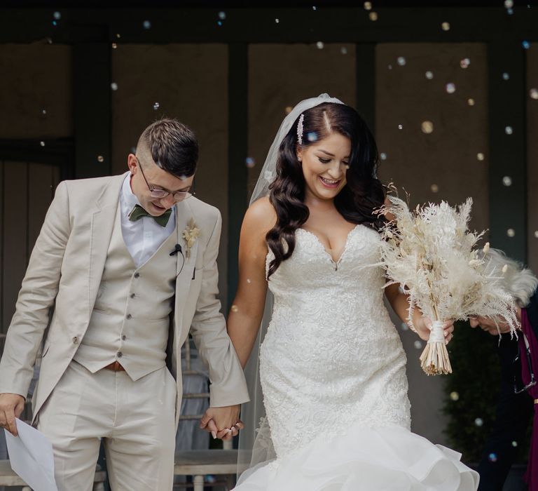 Bride in a lace fitted fishtail wedding dress with long wavy hair holding hands with her groom in a three-piece beige suit 