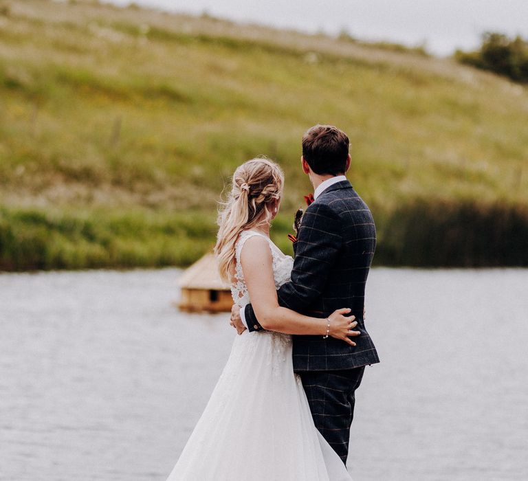 Bride & groom look across the lake and green hills as they wrap their arms around one another on their wedding day