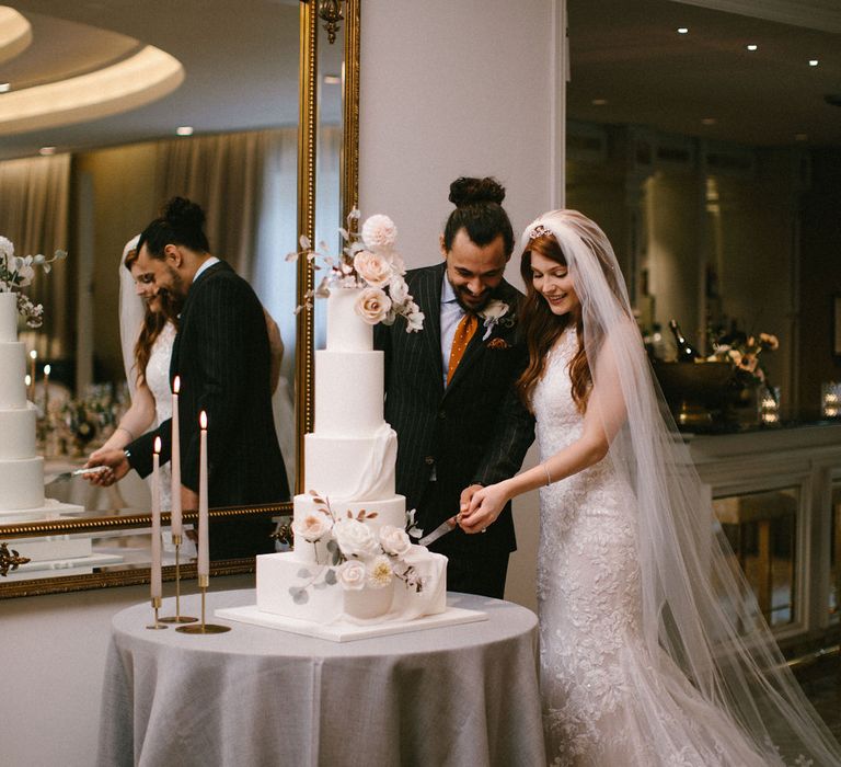 Bride in a lace Pronovias wedding dress cutting the tall iced wedding cake with her groom in a pinstripe suit and orange tie