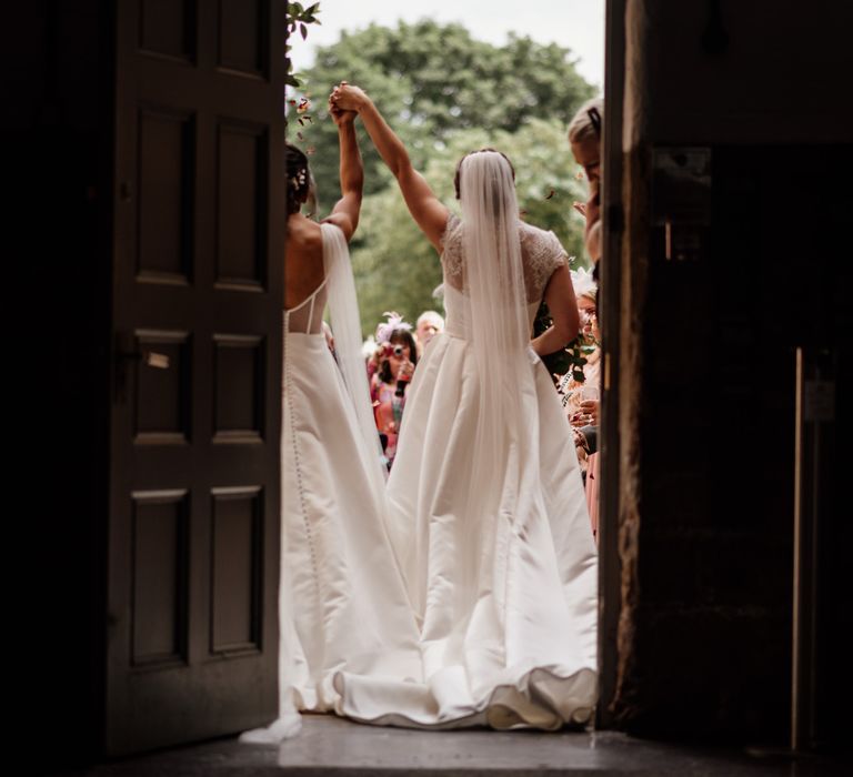 Bride in lace top wedding dress and veil holds arm in the air with bride in low back wedding dress with shoulder veils outside The West Mill Derby after wedding ceremony