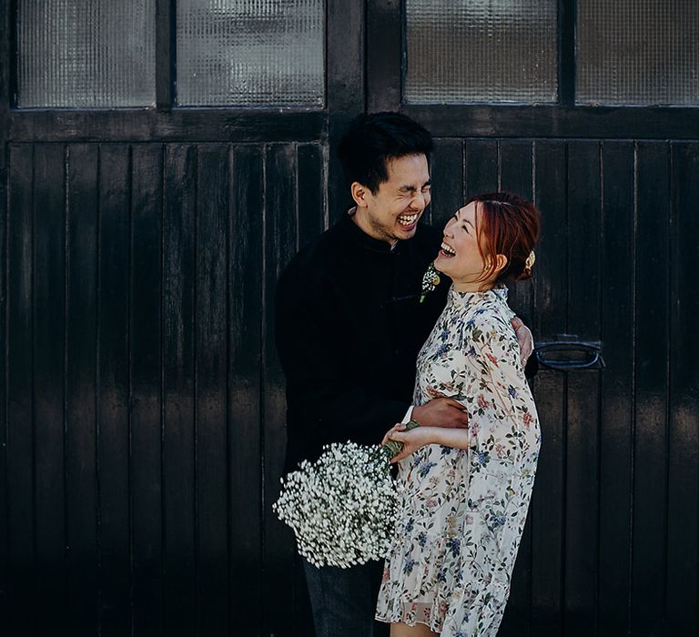 Portrait of the bride and groom smiling and laughing in front of a doorway with bride in a blue floral wedding dress with cape sleeves holding a gypsophila wedding bouquet 