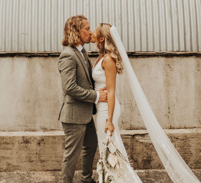 Bride in white Made With Love wedding dress and embroidered veil holding white and green dried bridal bouquet kisses groom in sage green suit and brown shoes outside after wedding ceremony at Anran Devon